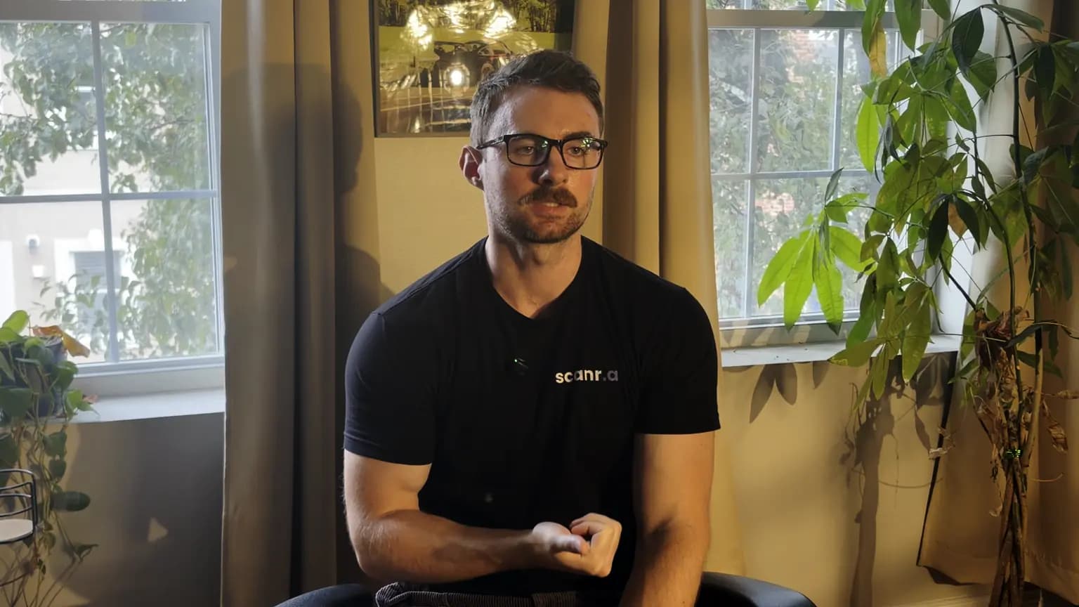 Eoin, the founder of Scanr.ai, in a black scanr.ai T-shirt, talking to camera in front of two windows and a tall houseplant.
